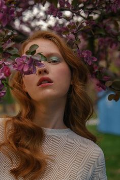 a woman with long red hair and green eyeshadow standing under a tree filled with purple flowers