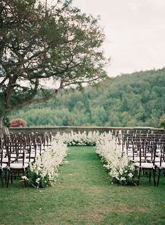 an outdoor ceremony set up with chairs and white flowers on the aisle, surrounded by greenery