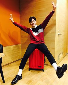 a man sitting on top of a red suitcase in front of a wooden wall with his hands up