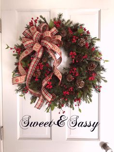 a christmas wreath with pine cones and red berries hanging on a white front door that says sweet & easy