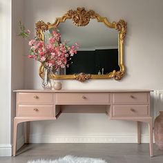 a pink desk with flowers on top and a gold framed mirror above it in a room