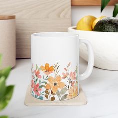 a white coffee mug sitting on top of a counter next to a bowl of fruit