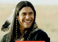 a man with long hair and feathers on his head smiles at the camera while holding a sign