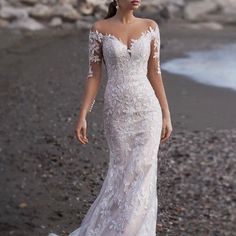 a woman is standing on the beach wearing a wedding dress with an open shoulder and long sleeves