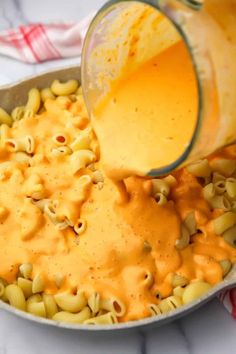 someone pouring orange sauce into a skillet filled with macaroni and cheese pasta