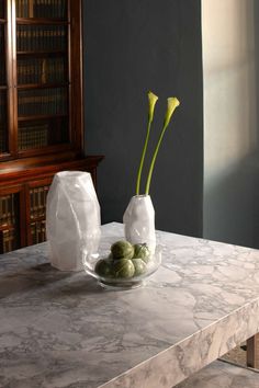 two white vases with flowers in them sitting on a marble table top next to bookshelves