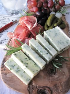 a wooden platter filled with cheese and crackers next to grapes, tomatoes, cucumbers and pickles