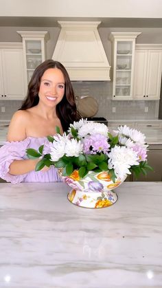 a woman sitting at a kitchen counter holding a vase with flowers in it and smiling