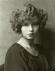 an old black and white photo of a woman with curly hair wearing a brown dress