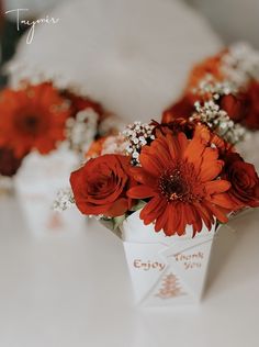 some red flowers are in a white cup on a table with other orange flowers and baby's breath