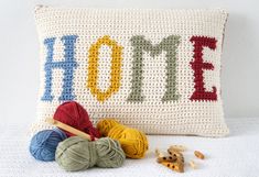 a crocheted pillow with the word home written on it next to yarn balls