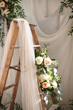 a wooden ladder with flowers and greenery on it next to a white curtained backdrop