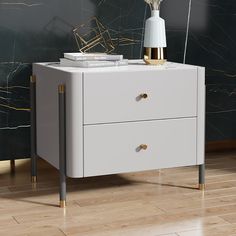 a white cabinet with two drawers and a gold vase on the top, in front of a black marble wall