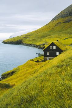 a small house on the side of a grassy hill next to water and grass covered hills