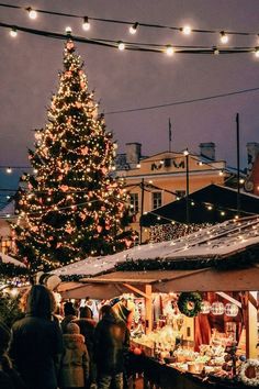 a christmas tree is lit up in the background as people walk by at an outdoor market