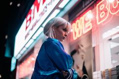 a woman with blonde hair standing in front of a building at night wearing a blue jacket