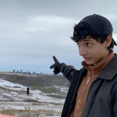 a young man pointing at something in the distance while wearing a black hat and leather jacket