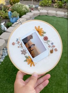 someone is holding up an embroidered photo frame with flowers and leaves on it in the grass