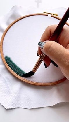 a woman's hand holding a pen and writing on a piece of paper in front of a cross - stitch hoop