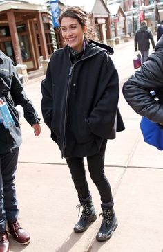 a woman in black jacket and boots standing on sidewalk with other people walking by her