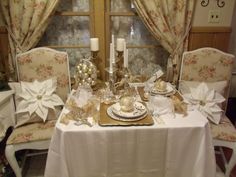 a table set for two with white and gold decorations on it, in front of a window