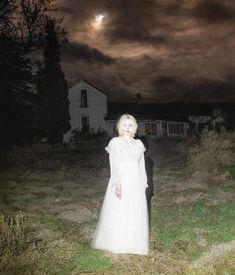 a woman in a white dress standing on grass next to a house with a full moon behind her