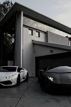 two sports cars are parked in front of a modern house, one is white and the other is black