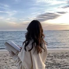 a woman standing on top of a sandy beach next to the ocean holding a book