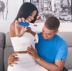 a pregnant woman brushing her husband's teeth with an electric toothbrush while sitting on a couch