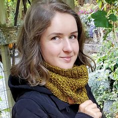 a woman wearing a black jacket and yellow scarf standing in front of some flowers with her hands on her hips