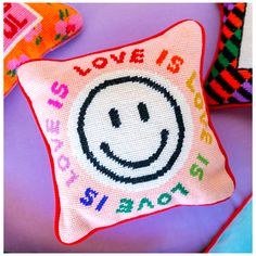 a cross stitch pillow with a smiley face on it and the words love is written in multicolored letters