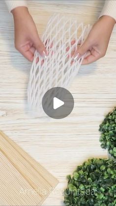 two hands holding white string on top of a wooden table next to green plants and wood planks