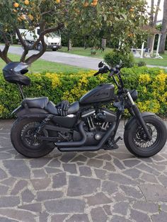 a black motorcycle parked on top of a stone road next to bushes and trees with oranges in the background