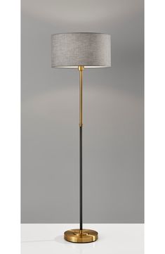 a floor lamp with a white shade on top and a gold base, in front of a gray wall