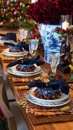 the table is set for christmas dinner with blue and white plates, silverware, red flowers and greenery