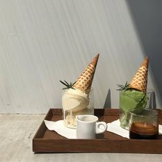 two ice cream cones sitting on top of a wooden tray next to cups and saucers