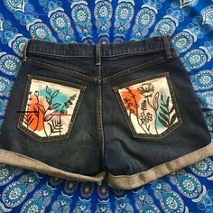 a pair of jean shorts with flowers painted on the front and back pockets, sitting on top of a paisley print cloth