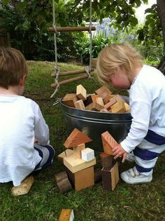 two young boys playing with wooden blocks in a yard next to a metal bucket full of wood