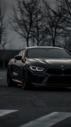 the front end of a black car parked in a parking lot with trees behind it