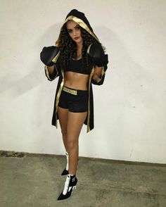 a woman in black and gold outfit holding a pair of boxing gloves while standing next to a wall