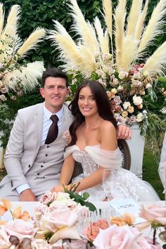 a man and woman sitting at a table with flowers in front of them, posing for the camera