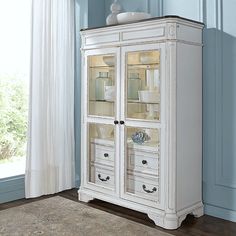 a white china cabinet with glass doors in a blue walled room next to a window