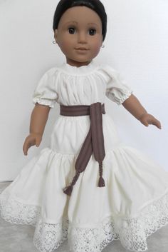 an american girl doll wearing a white dress and brown ribbon around her waist, standing on a gray surface