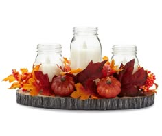 three glass jars filled with candles sitting on top of a wooden tray surrounded by fall leaves and pumpkins