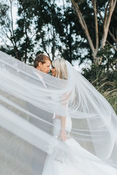 a bride and groom kissing under a veil in front of some tall grass with trees behind them