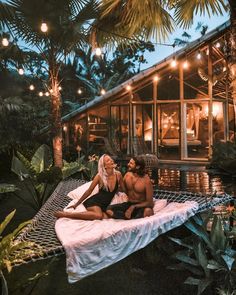 two people sitting on a hammock in front of a house with lights strung from the roof