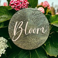 a wooden sign that says bloom on top of some leaves and flowers in the background