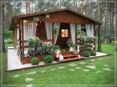 a small wooden cabin in the middle of a forest with flowers and plants on the porch