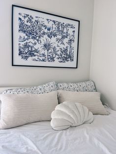 a white bed topped with lots of pillows under a framed art piece on the wall