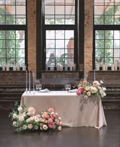 a table with flowers and candles on it in front of two large windows at the end of a room